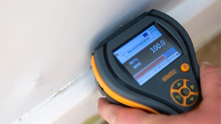 A moisture meter being held against a wall with signs of damp. The reading shows 100% saturation.