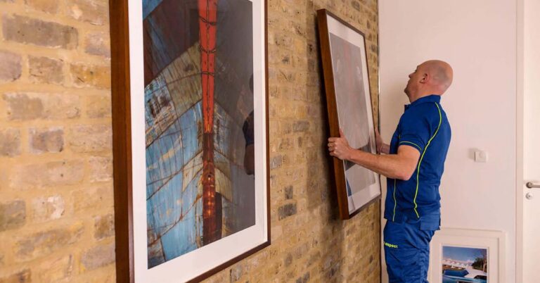 An Aspect handyman hanging a large framed picture on a wall next to another picture.