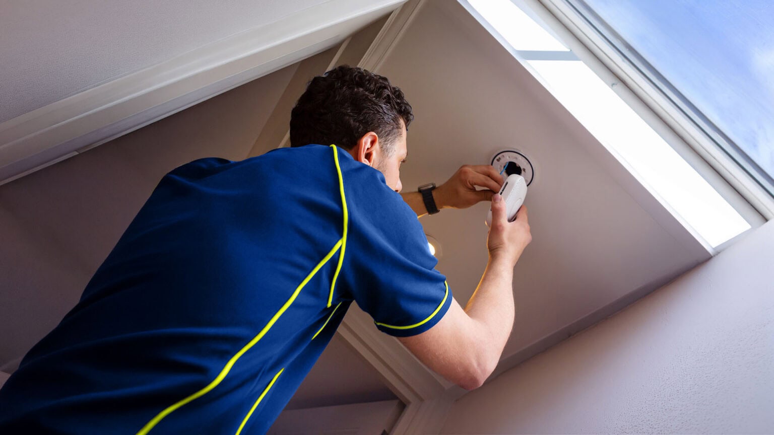 An Aspect electrician wiring a smoke alarm on a ceiling next to a roof window.