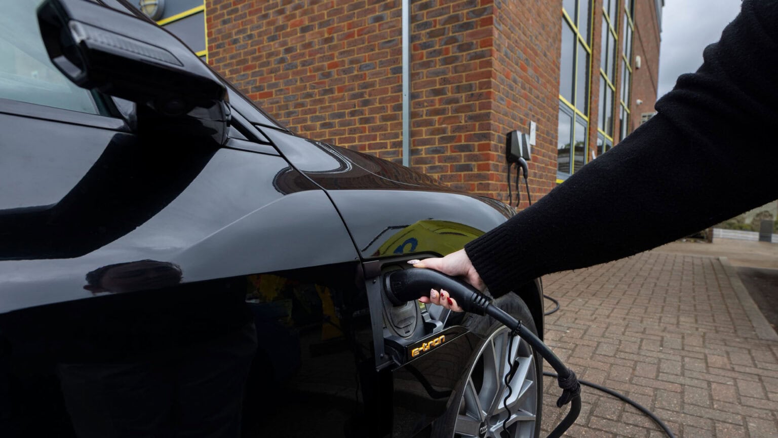 A person's hand plugging their electric car into a charger unit that is mounted on the wall behind.
