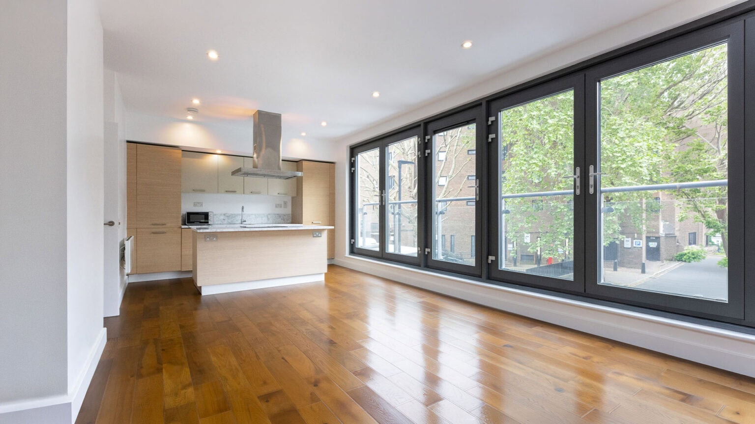 A newly refurbished open-plan living space with a kitchen at one end, a polished hardwood floor, and floor to ceiling metal frame windows along one side.